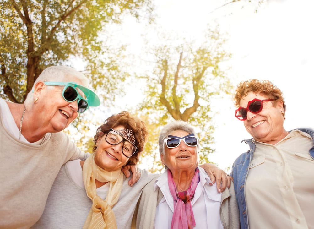 Group of older women wearing fun sunglasses pose with their arms around each other  